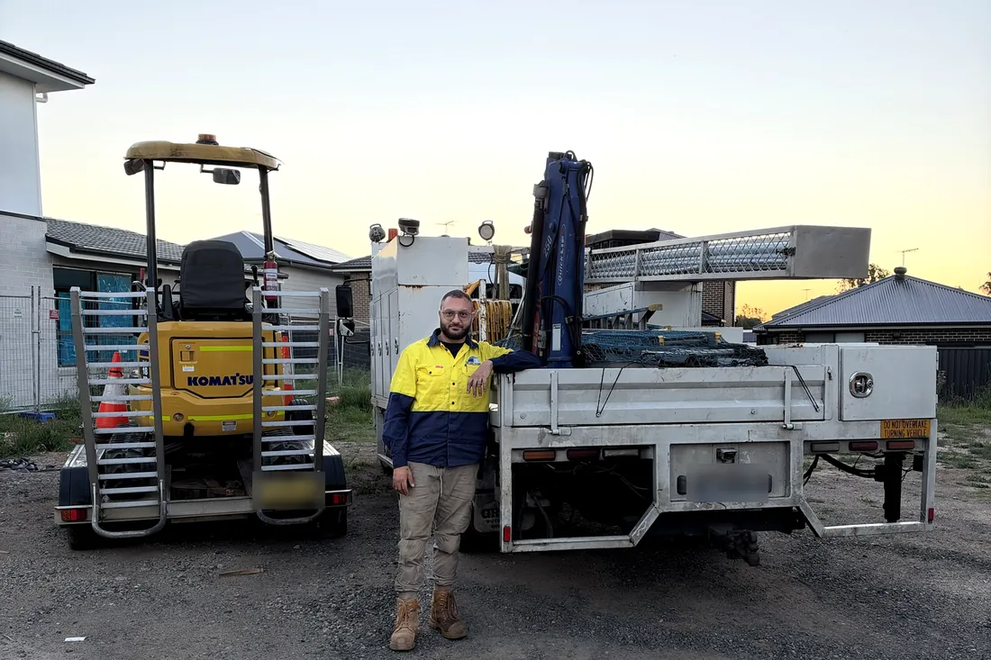 Paul, owner of Monarch Civil & Plumbing, standing between the Komatsu PC18 excavator on its trailer and the crane truck